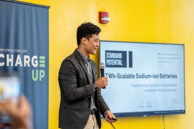 Young male holding microphone presents sodium-ion battery technology solution at ChargeUp final pitch event in Binghamton, NY