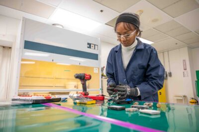 Young female wearing personal protection equipment stands at worktable with tools and materials, taking apart used, discharged batteries.